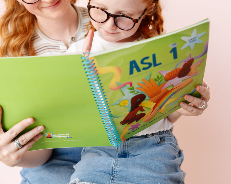 A child wearing glasses and a striped shirt holds an open spiral-bound ASL 1 book with colorful illustrations, while an adult with red hair sits behind them against a soft pink background.